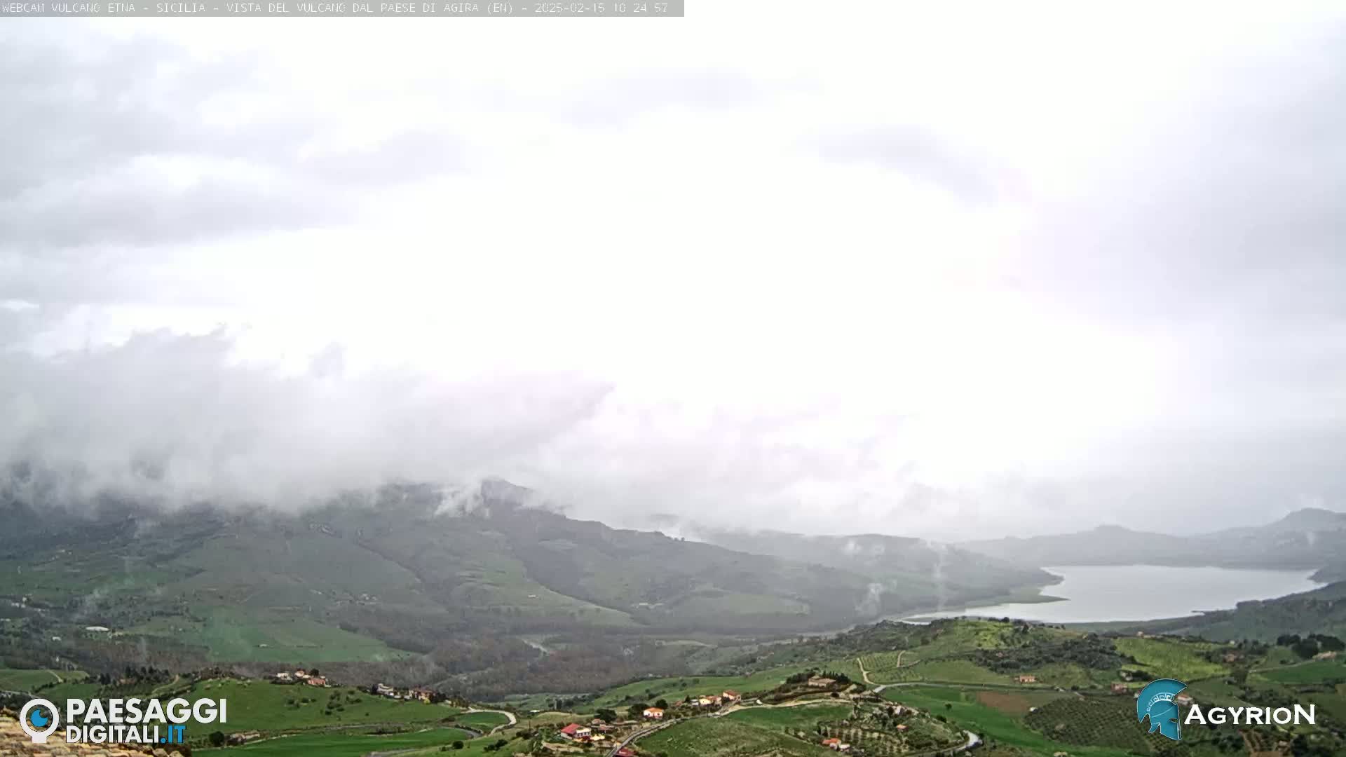 A cloudy day reveals a valley with rolling green hills, farmland, and a lake, partially obscured by low-hanging clouds.
