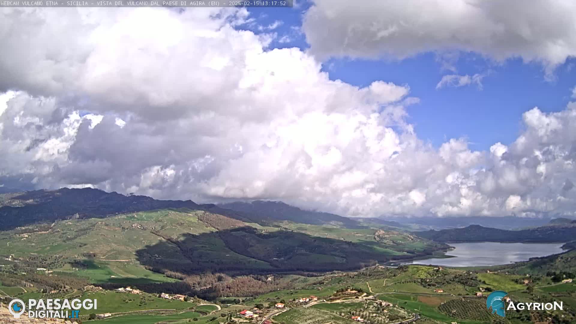 A partly cloudy day reveals a sprawling valley landscape with a lake, rolling hills, and small villages, all under a sky filled with fluffy white and gray clouds.