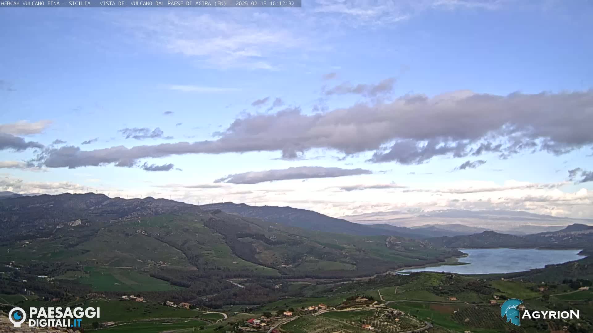 A partly cloudy sky overlooks a vast, hilly landscape with a lake in the distance.