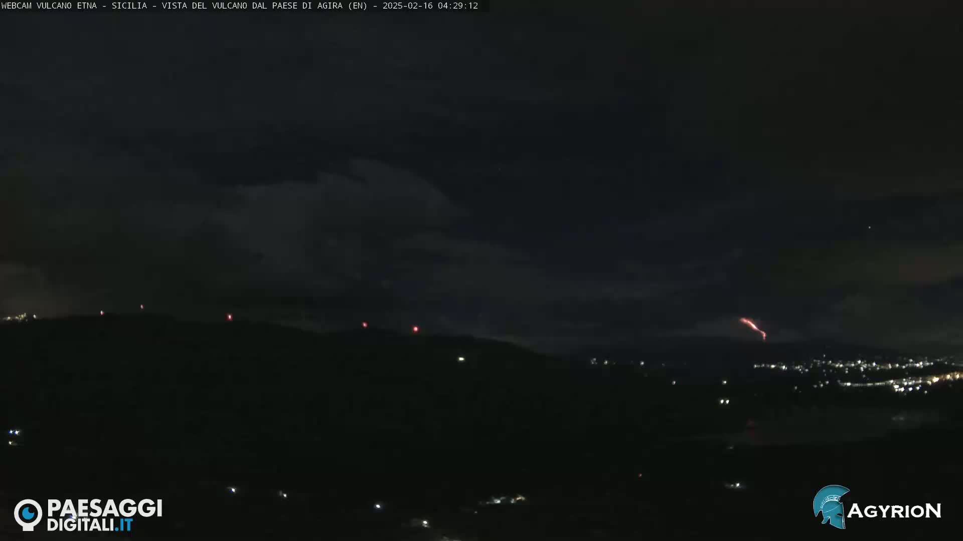 A nighttime, possibly cloudy, long shot of a dark landscape with a faint reddish glow emanating from a distant mountain range, and a few scattered lights visible below.