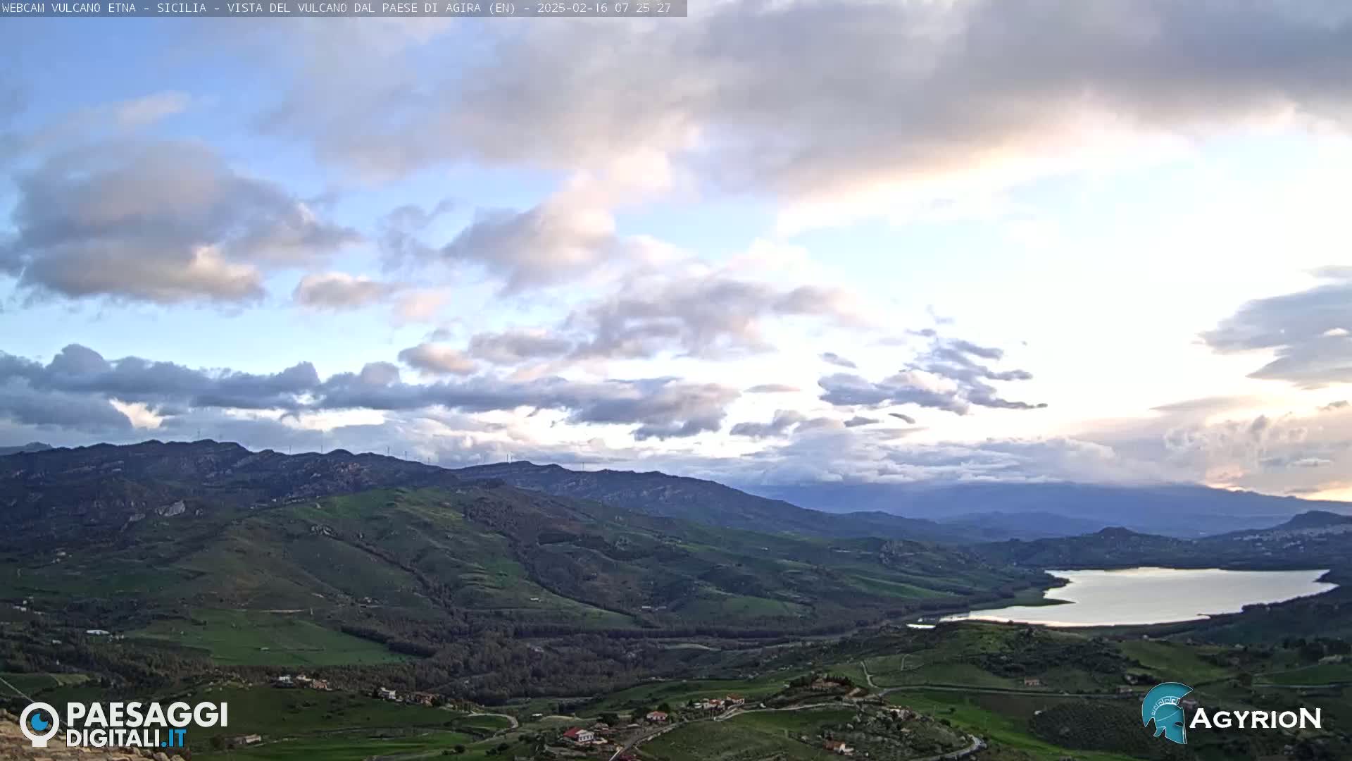 A partly cloudy sky overlooks a vast, rolling green landscape with a lake and distant mountains.