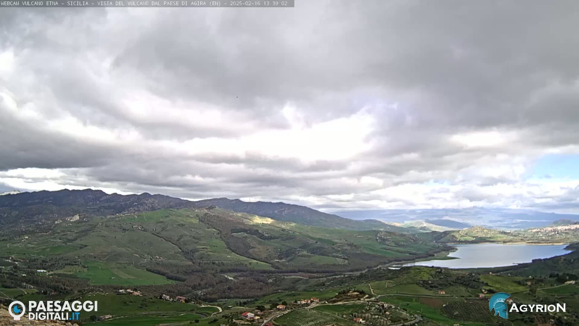 A wide, hilly landscape under a mostly cloudy sky shows a large lake nestled in a valley.