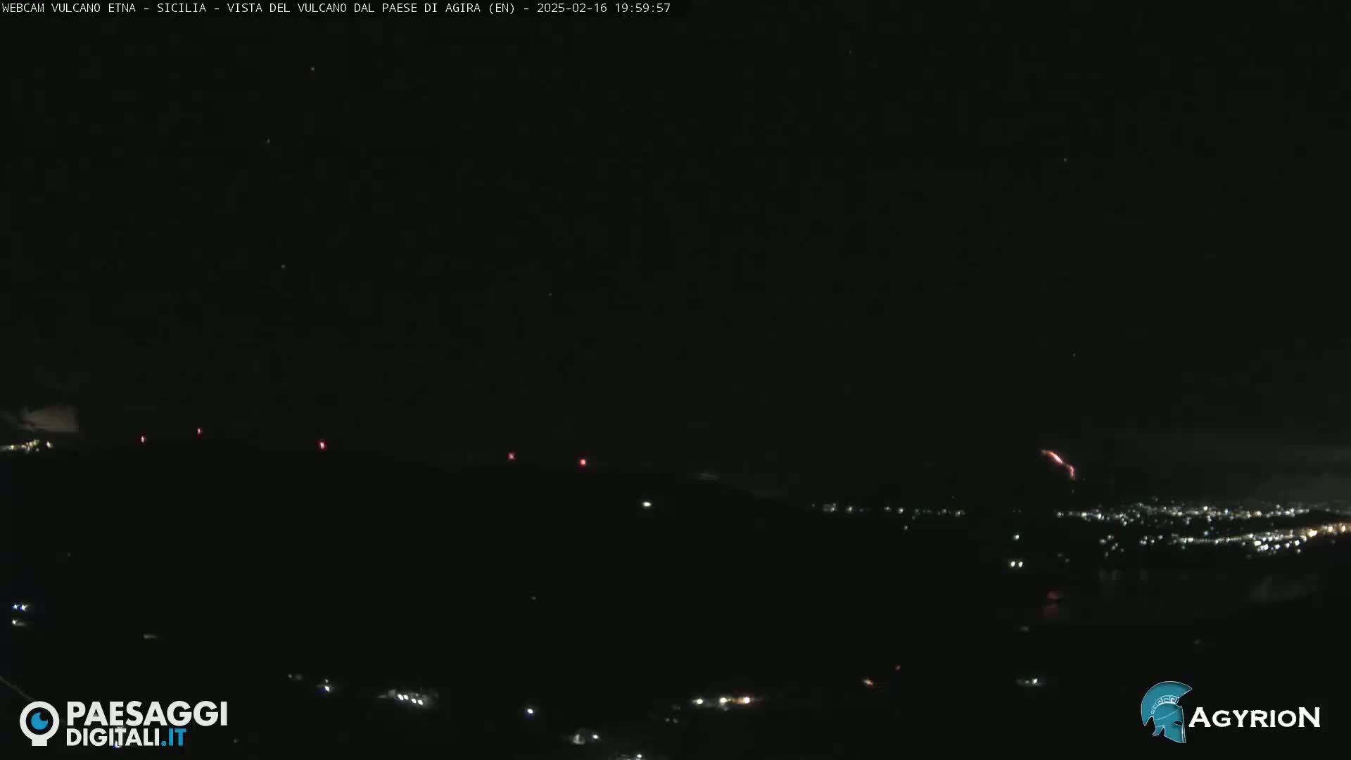 A nighttime long shot shows a dark landscape with a distant city illuminated by lights and a faint reddish glow near the horizon, possibly indicating volcanic activity.