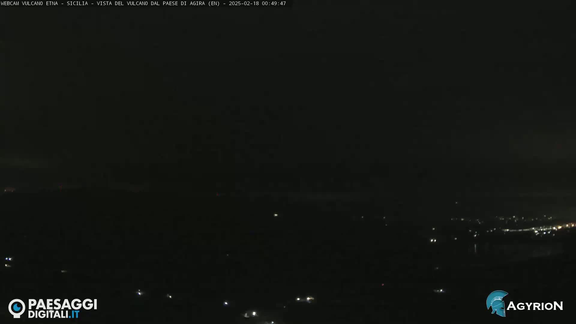 A nighttime long shot of dimly lit hills and a town in the distance under a dark, cloudy sky.