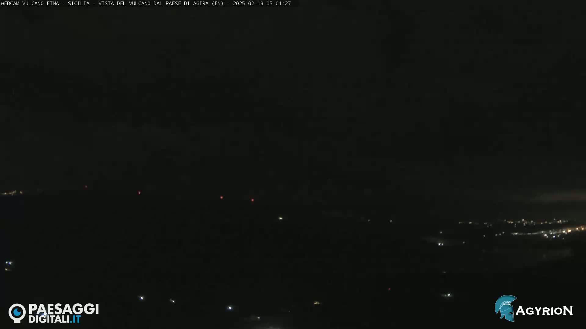 A nighttime long shot shows a dark landscape with a few faint lights scattered across the bottom and a few reddish lights further up, under a dark sky.