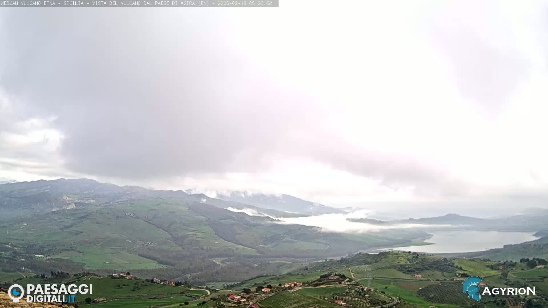 A misty, overcast day reveals a valley with rolling green hills, farmland, and a lake nestled among the mountains.