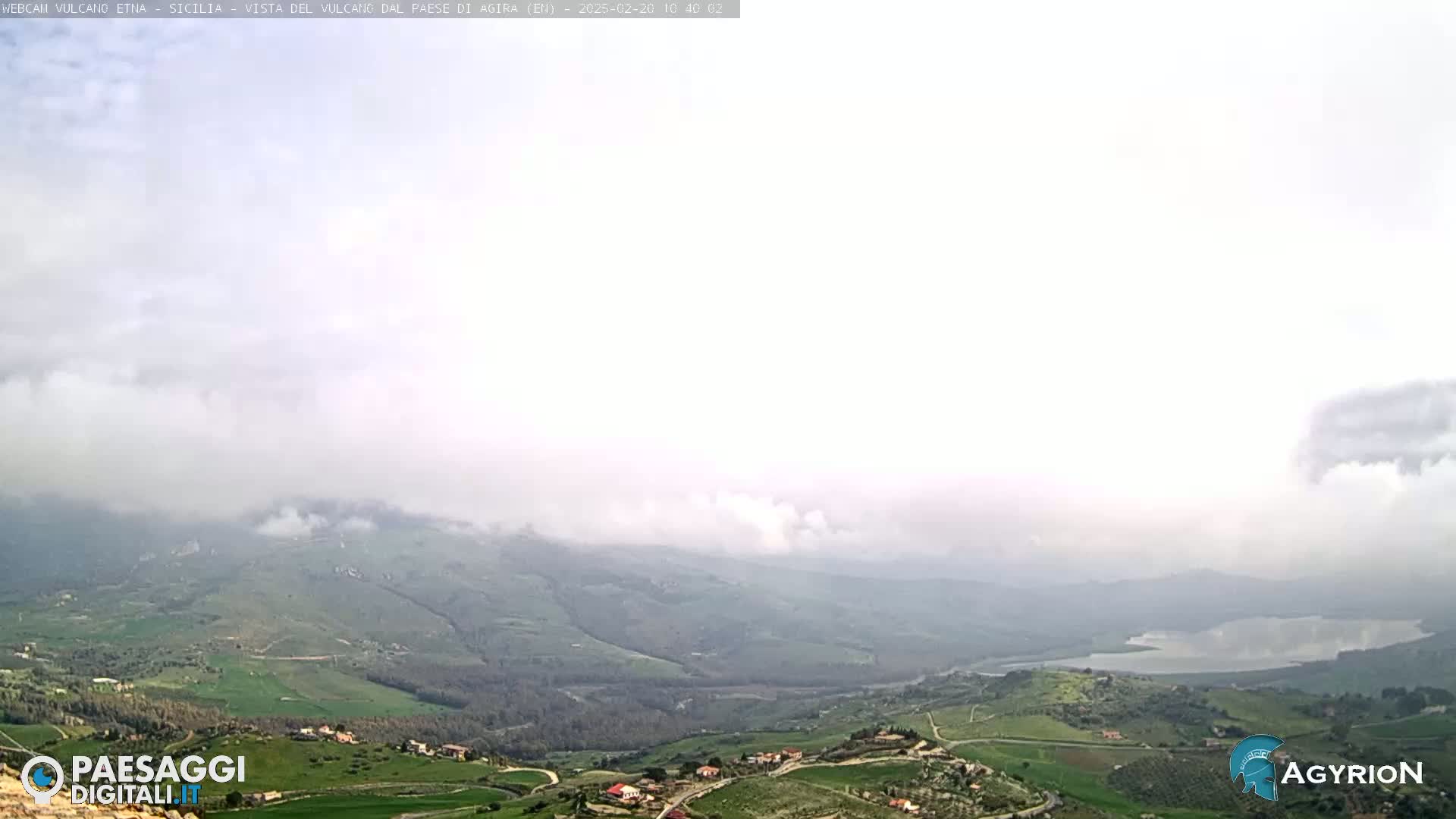 A hazy, overcast day reveals a valley with rolling green hills, farmlands, a lake, and distant mountains partially obscured by clouds.