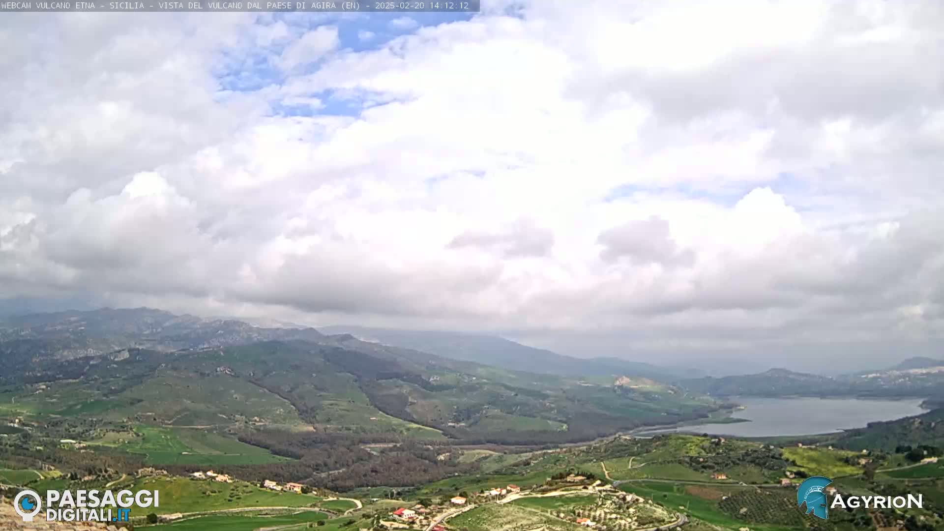 A partly cloudy day overlooks a vast, verdant valley containing a lake and small settlements, nestled between rolling hills.