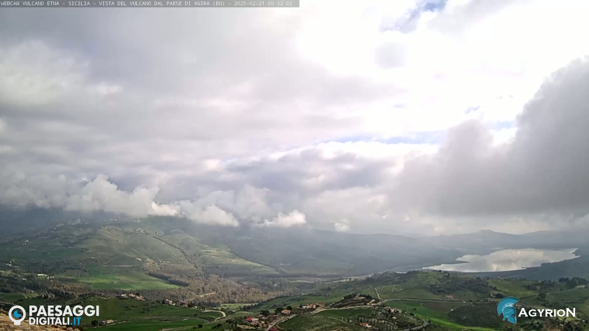A panoramic view of a hilly landscape with a lake, under a mostly cloudy sky.