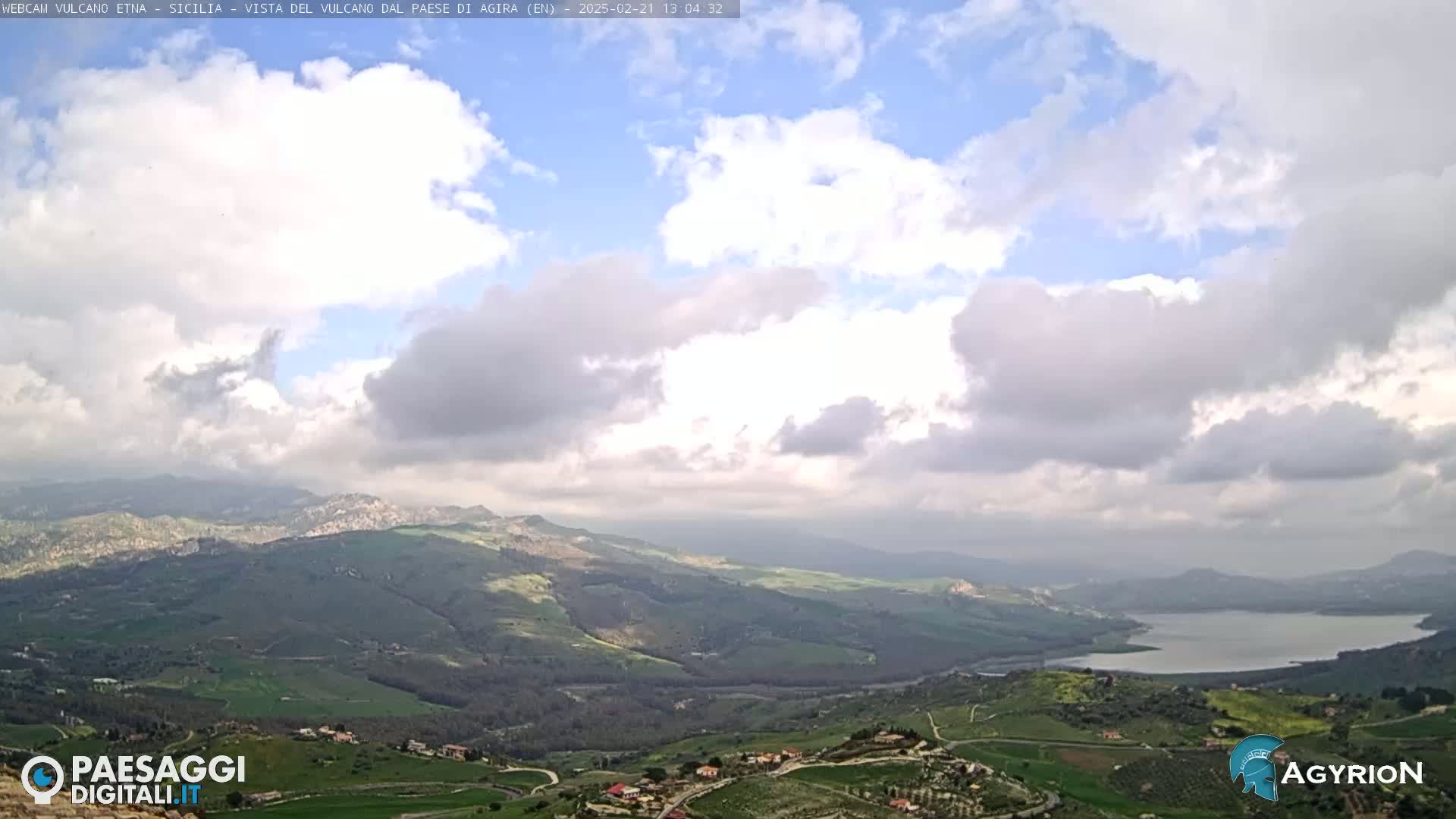 A partly cloudy sky overlooks a vast, hilly landscape with a lake visible in the distance.