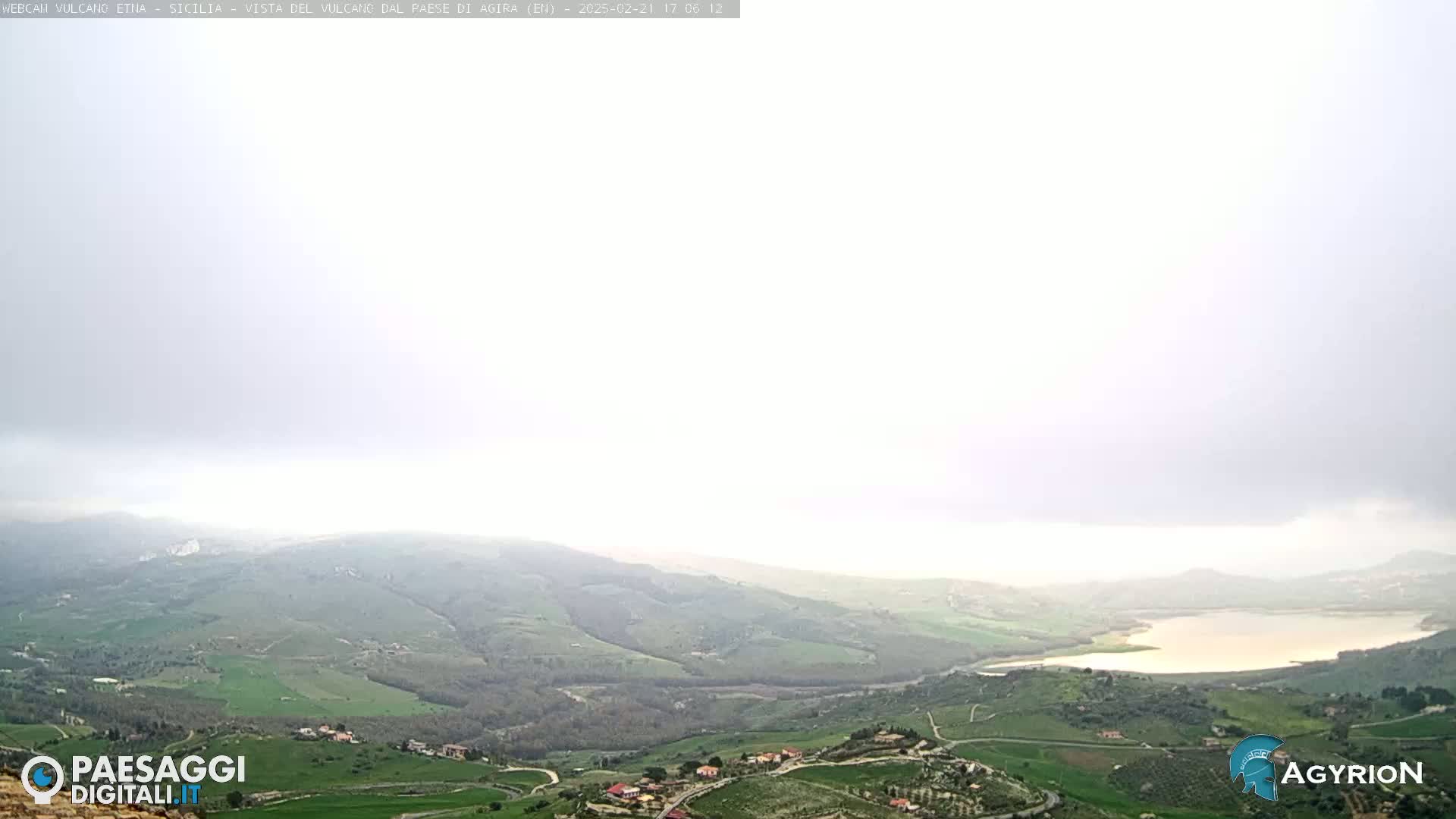 A hazy, overcast view shows a vast, rolling green landscape with a lake nestled in a valley, and small settlements scattered throughout.