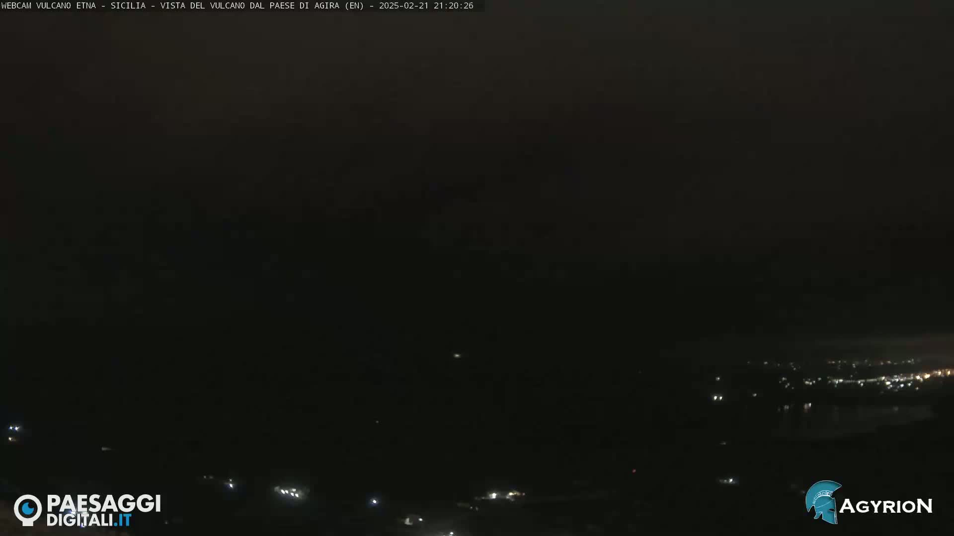 A nighttime long shot shows a dark, overcast sky over a dimly lit town by a body of water.