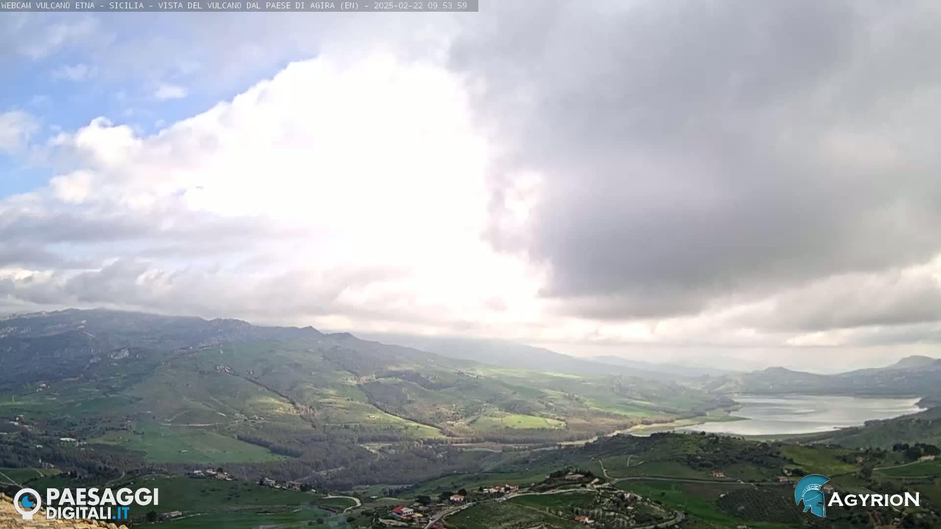 A partly cloudy sky overlooks a vast, rolling green landscape with a lake nestled in the valley below.