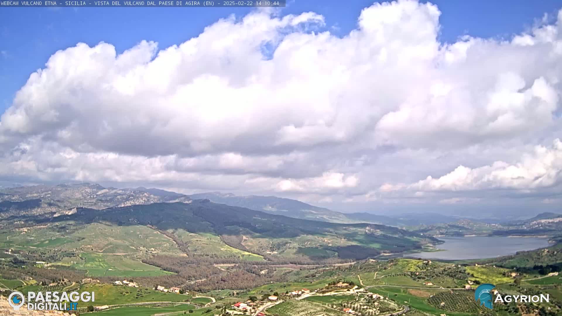 A partly cloudy sky overlooks a valley with rolling hills, farmland, and a lake in the distance.