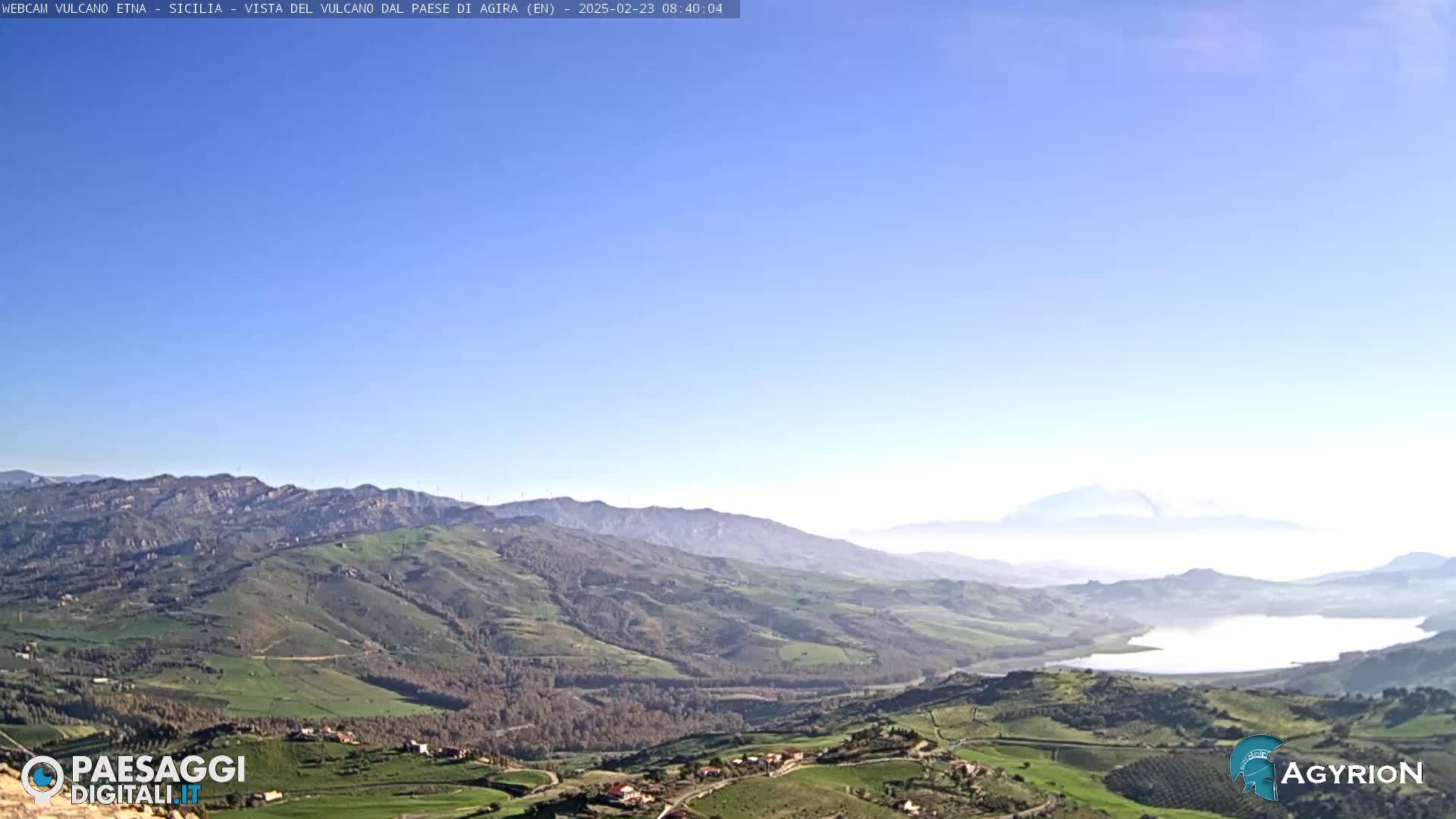 A clear, sunny day reveals a vast, rolling landscape of green hills and valleys, with a lake nestled among them and a distant, hazy mountain range.