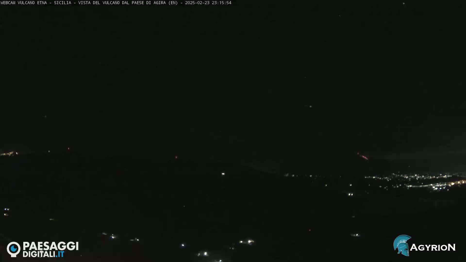 A nighttime long shot of a dark, hilly landscape with a few scattered lights, possibly a town, in the distance, and faint reddish glows near the horizon.