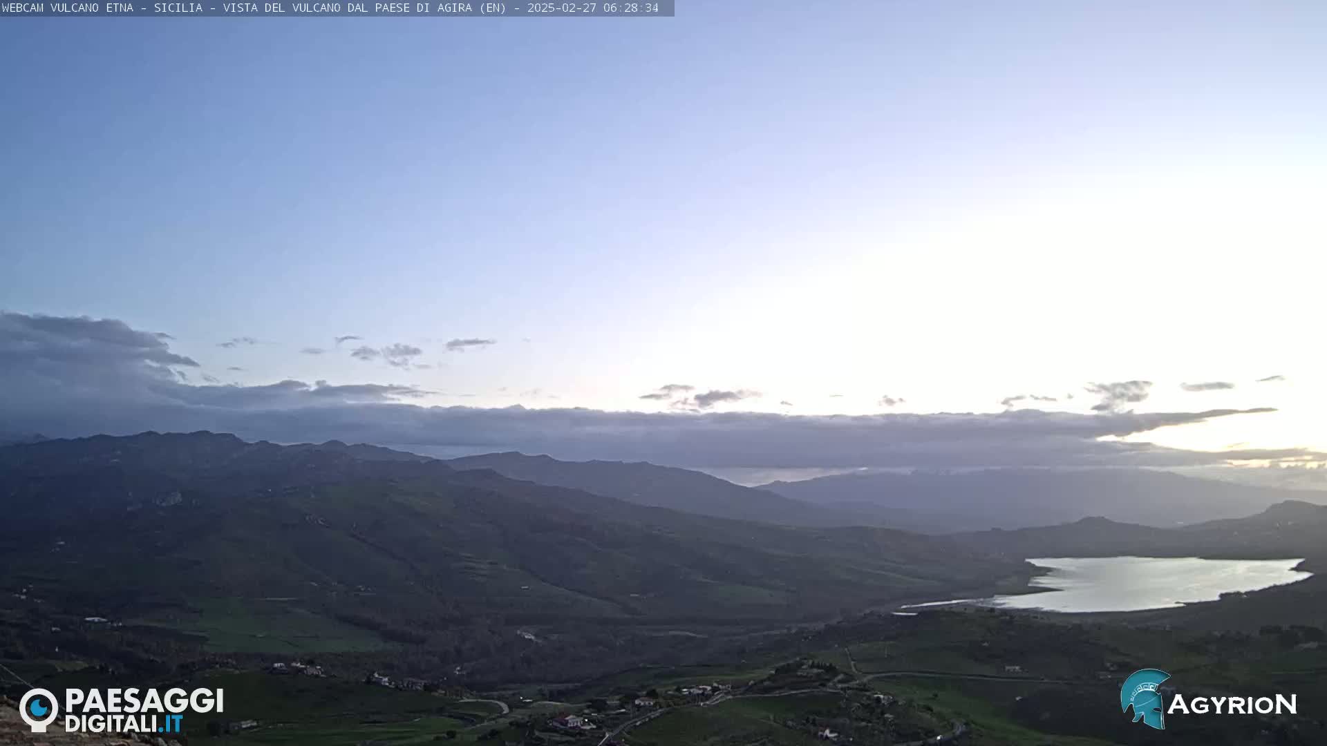 A sunrise view of rolling hills and a lake, with a hazy sky containing thin clouds.