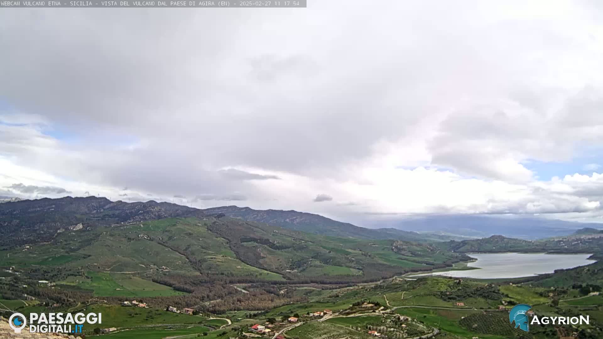 A hilly landscape with green fields and a lake is visible under a mostly cloudy sky.
