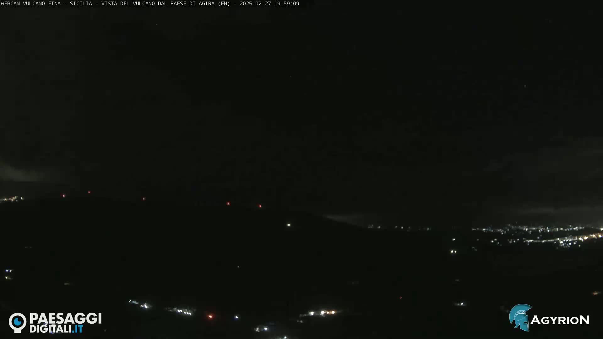 A nighttime, long shot of a dark, hilly landscape with a few scattered lights visible in the distance under a mostly clear sky.