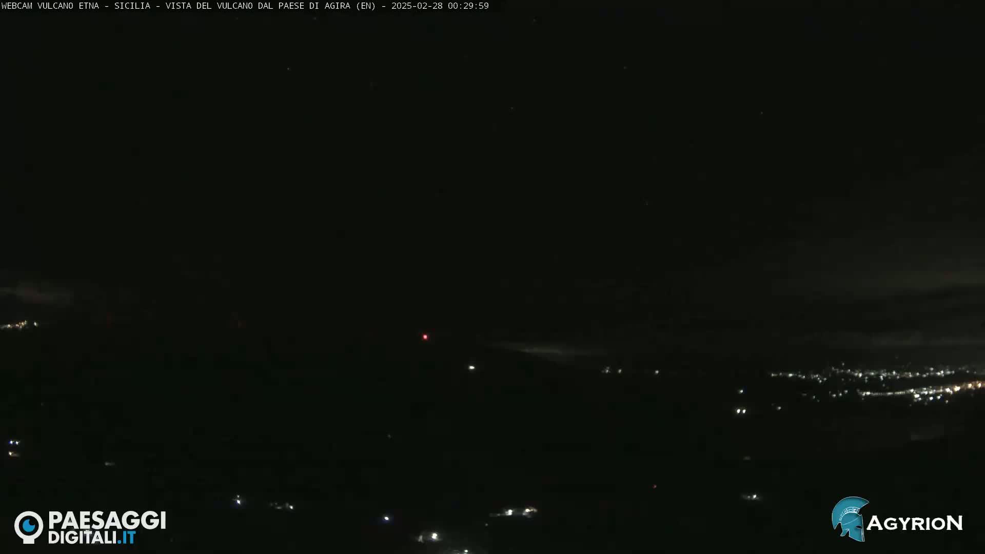 A dark, nighttime view of a distant city illuminated by scattered lights, under a mostly clear sky with a faint reddish glow possibly from a distant light source.