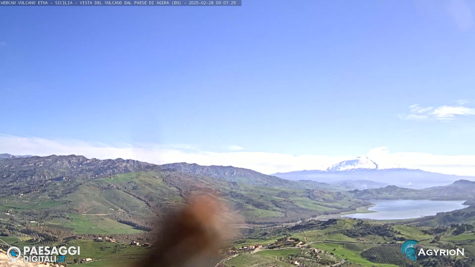 A snow-capped volcano is visible in the distance across a valley of rolling green hills and a lake under a clear blue sky.