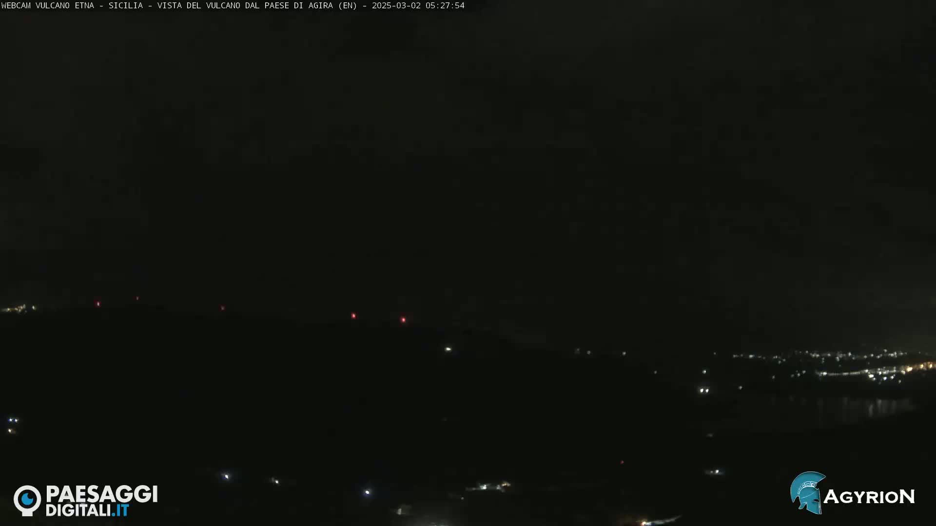 A nighttime long shot shows a dark landscape with a few scattered lights, possibly a town in the distance, under a mostly clear, dark sky.