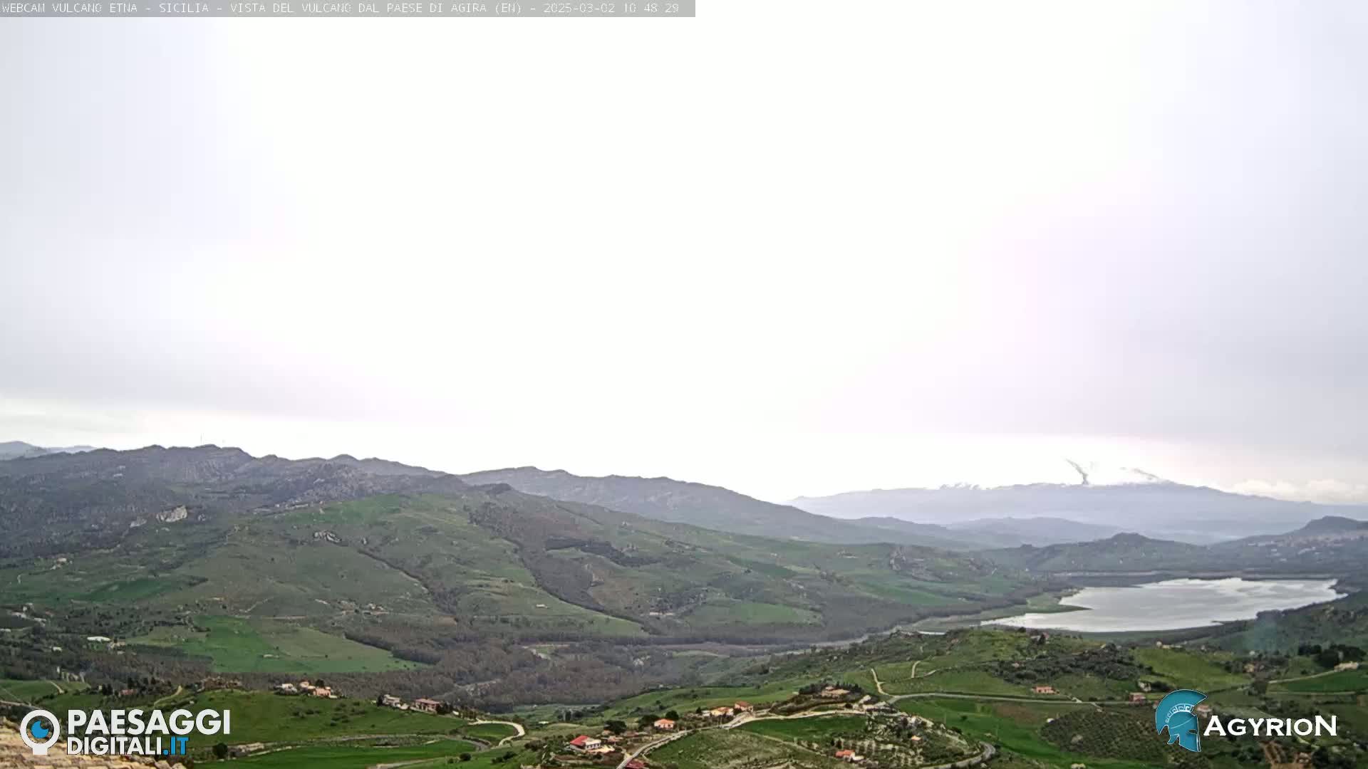 A distant, snow-capped mountain range is visible under an overcast sky, overlooking a valley of rolling green hills, farmland, and a lake.