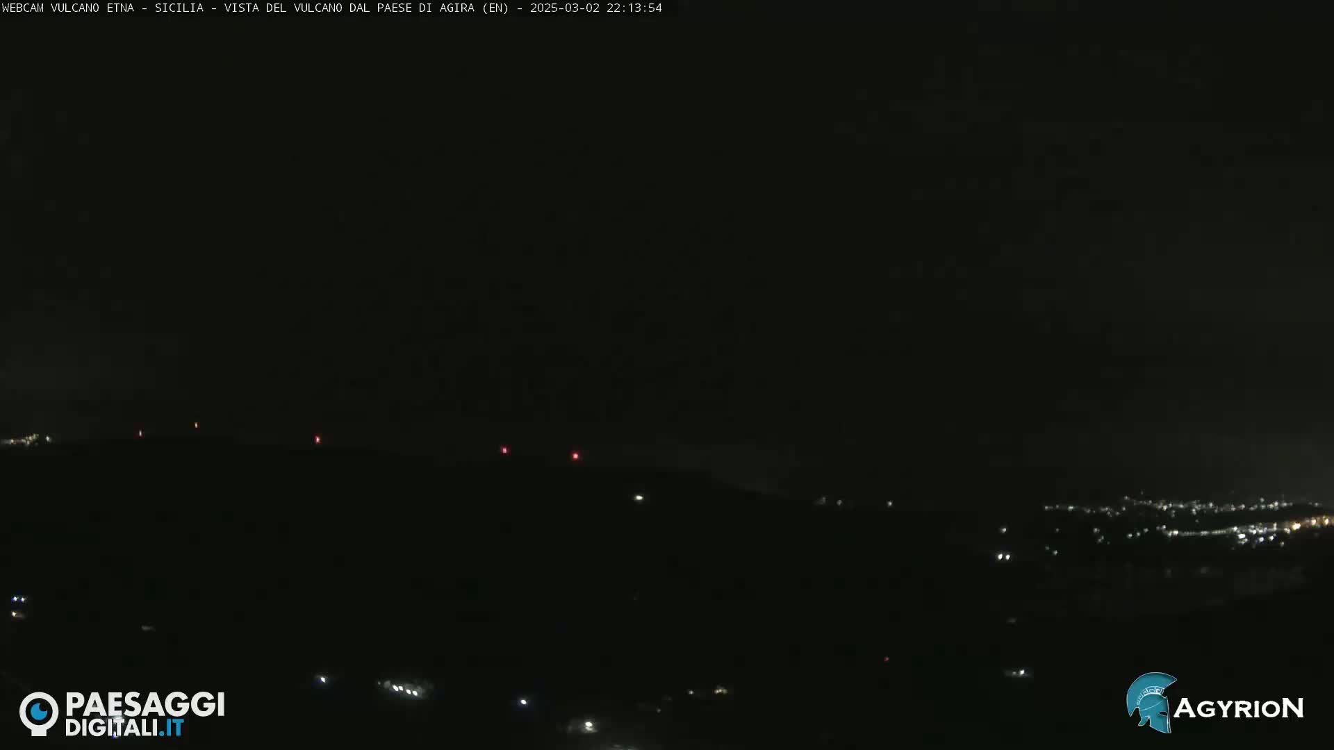 A nighttime long shot of a dark, hilly landscape with a few faint lights visible in the distance under a clear, dark sky.