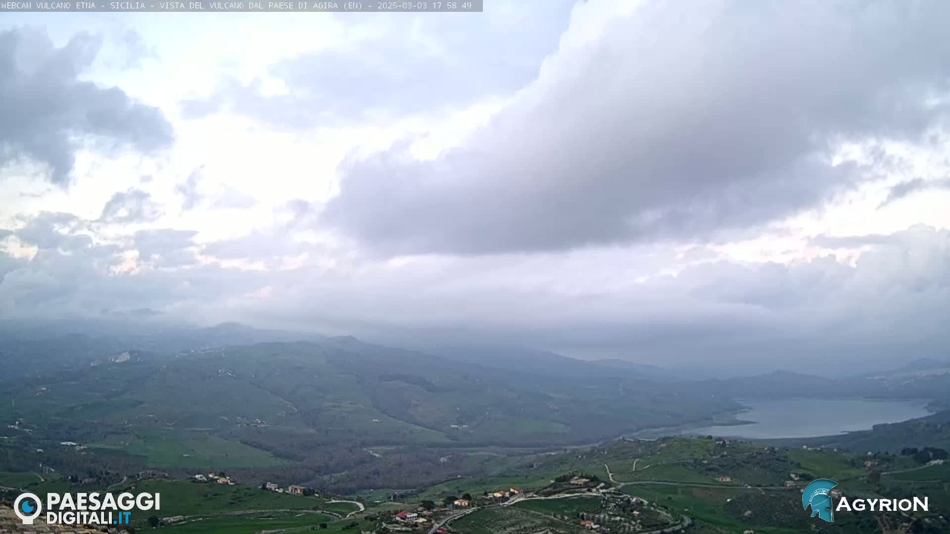 A sprawling, hilly landscape with a lake is visible under a mostly cloudy sky.