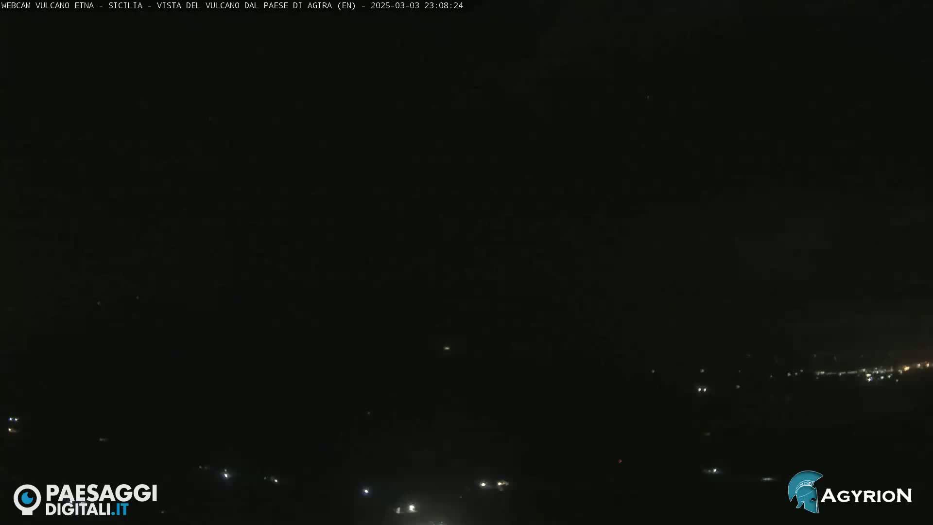 A nighttime long shot of a dark landscape with a few distant lights visible.