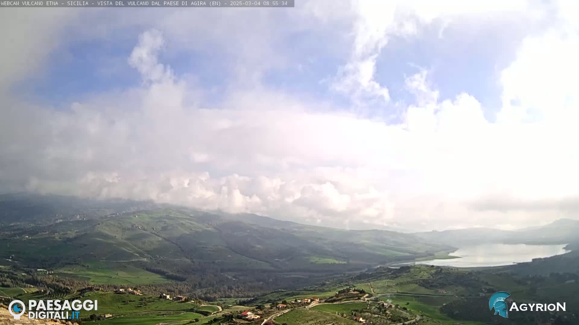 A partly cloudy sky overlooks a vast, green, hilly landscape containing a lake.