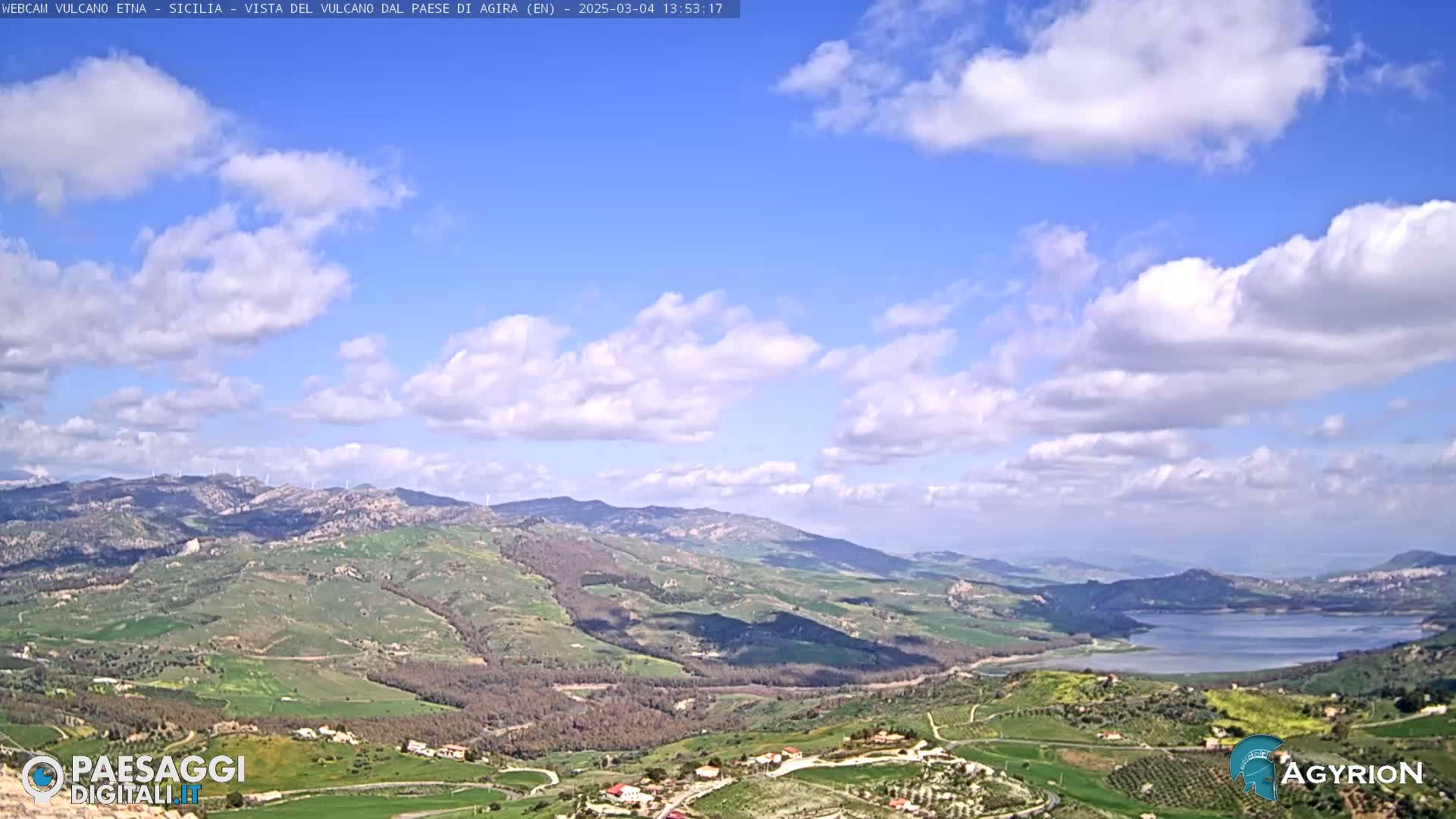 A partly cloudy blue sky overlooks a vast, hilly landscape with a lake visible in the distance.