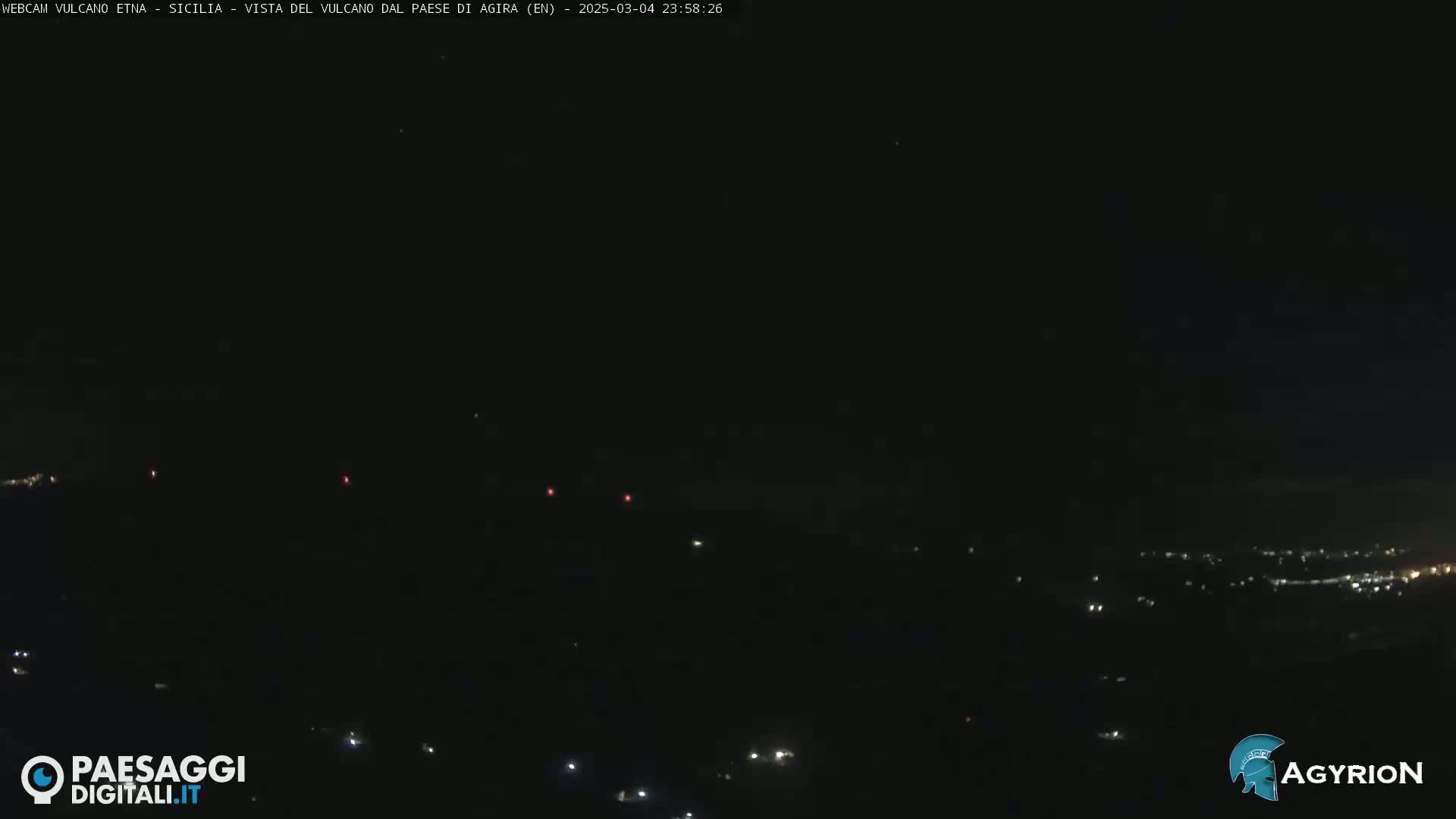 A nighttime, long-range view of a dark landscape with scattered lights, possibly a town, in the distance.