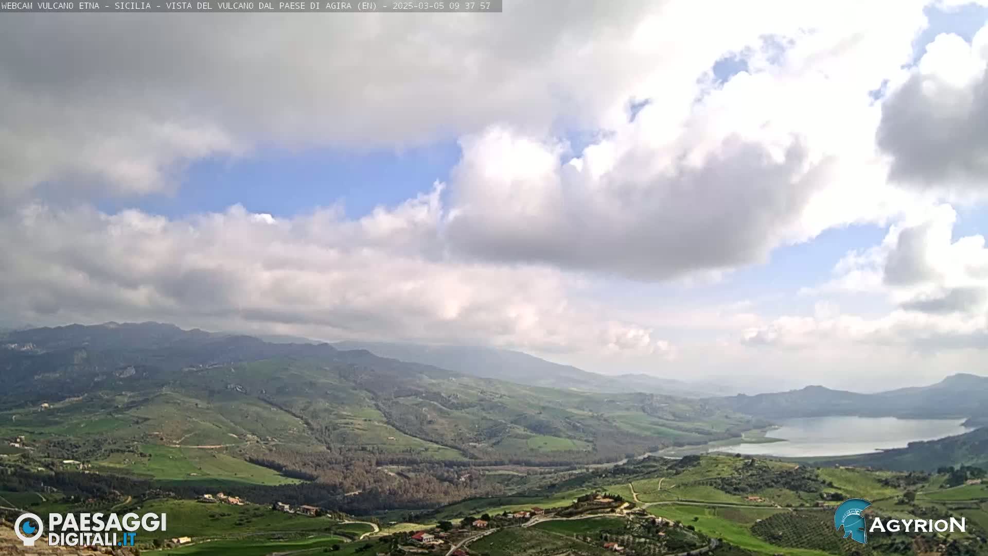 A partly cloudy sky overlooks a vast, verdant valley containing a lake and rolling hills.