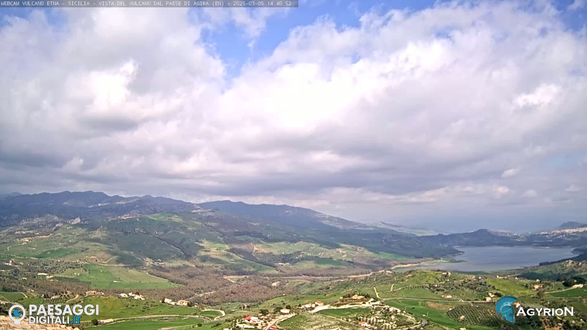 A partly cloudy sky overlooks a verdant valley with a lake and distant mountains.