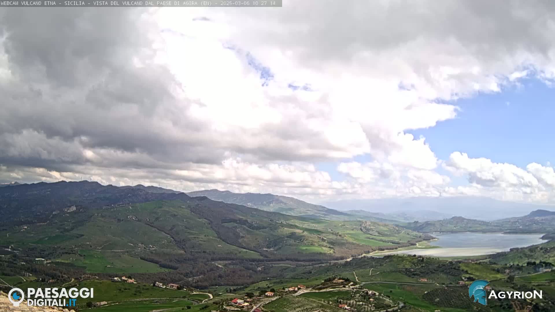 A partly cloudy day reveals a sprawling landscape of rolling green hills and a lake, with distant mountains under a mostly cloudy sky.