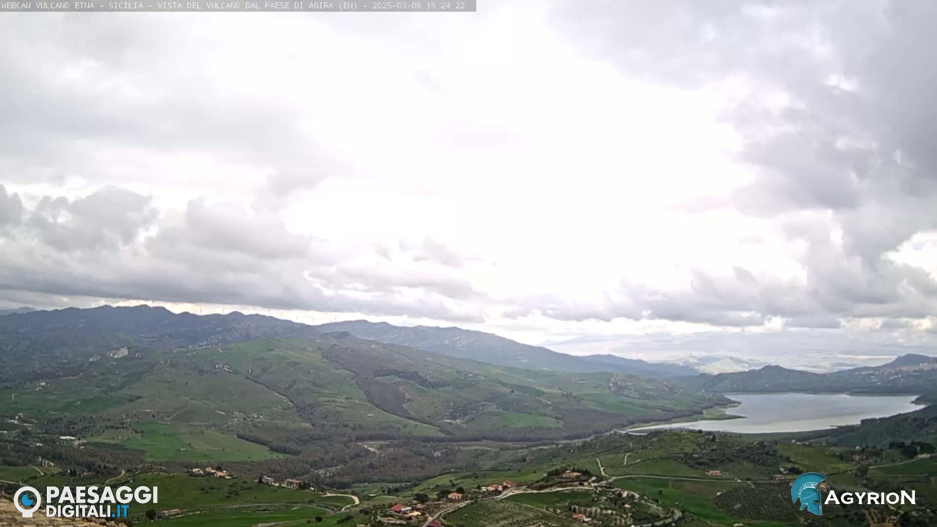 A mostly cloudy sky overlooks a hilly, green landscape with a lake in the distance.
