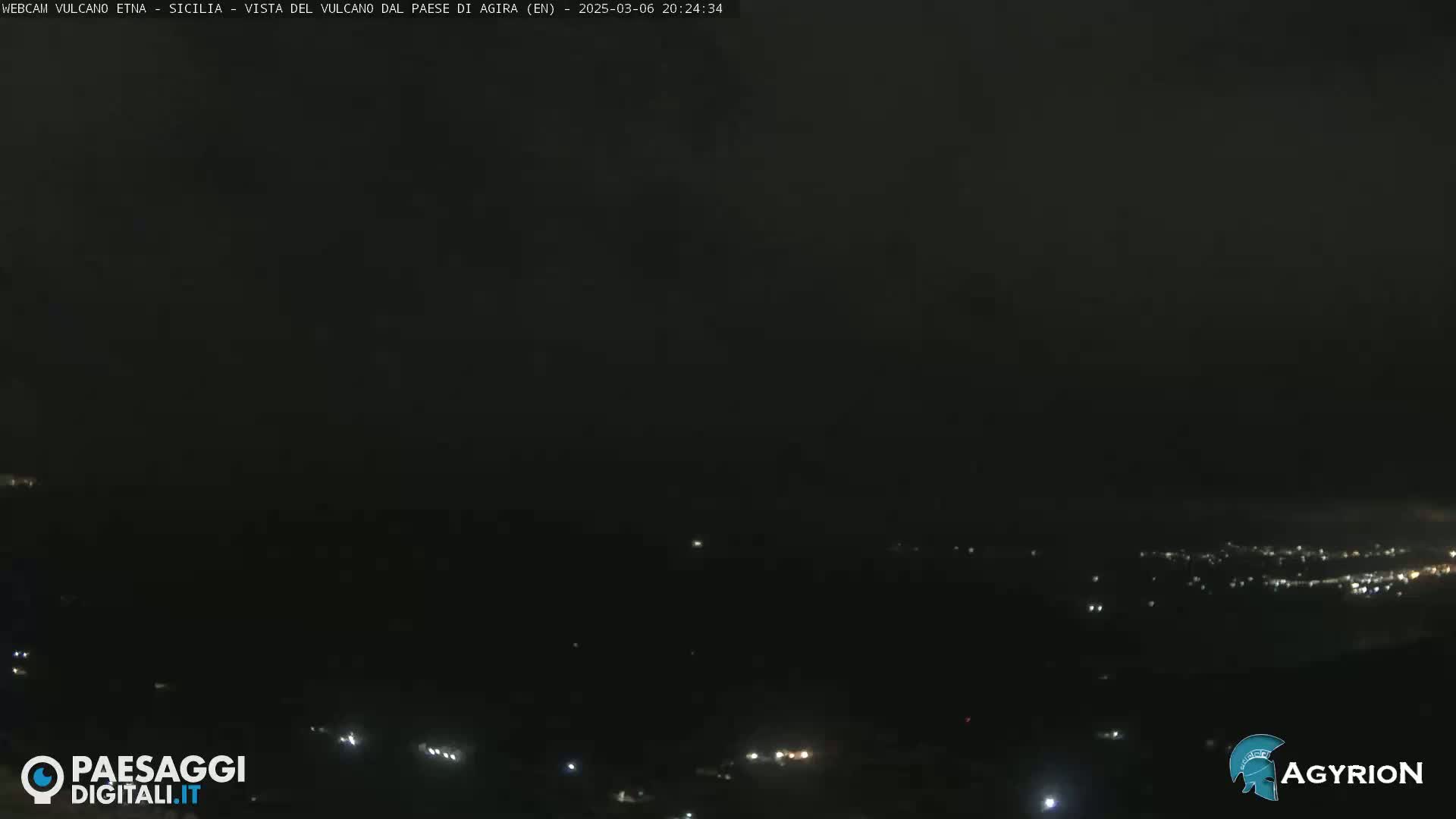 A nighttime long shot of a dark, hilly landscape with scattered lights from a town below, under a mostly clear, dark sky.