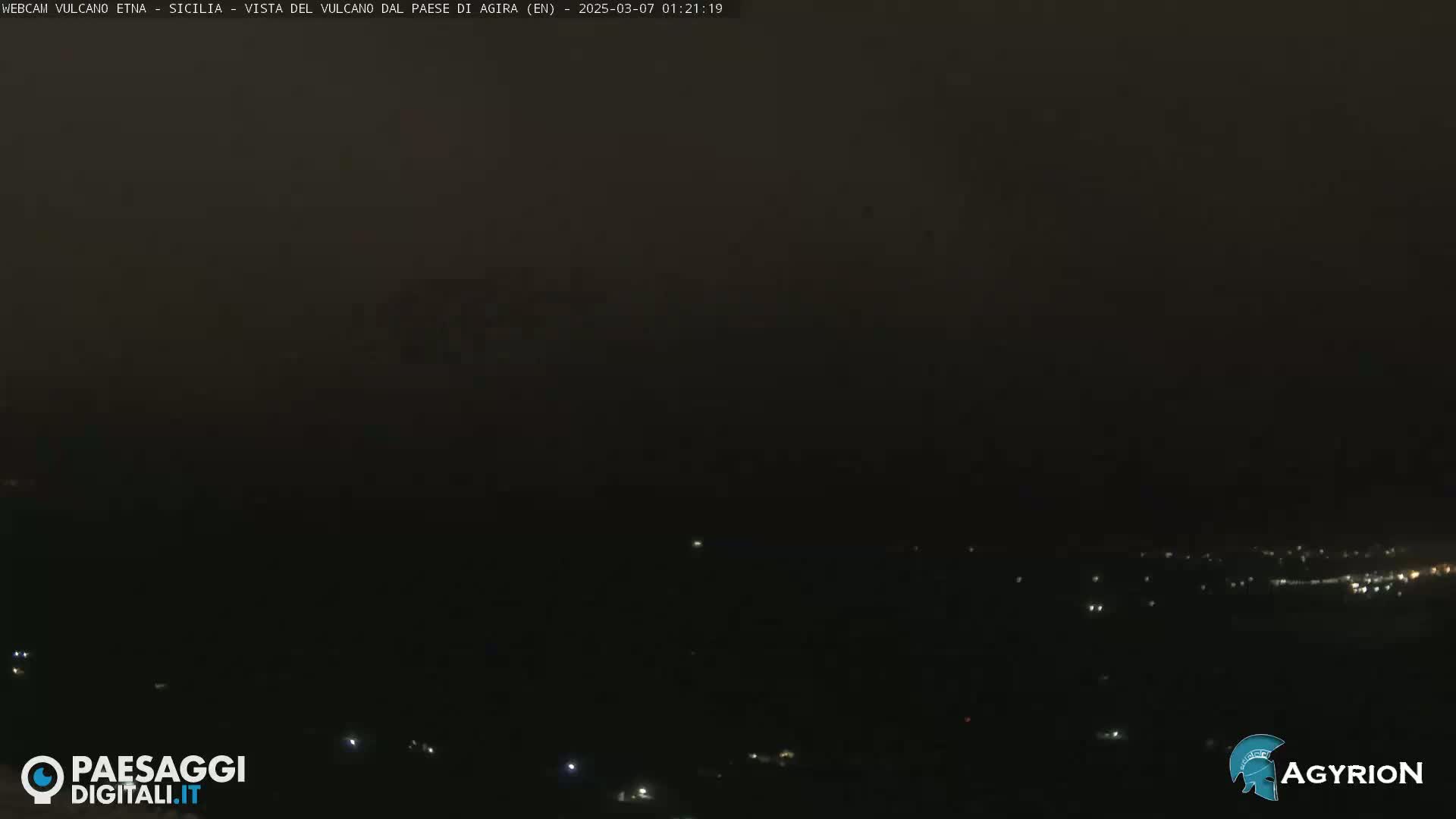 A dark, nighttime view of a distant town with scattered lights under a hazy, dark sky.