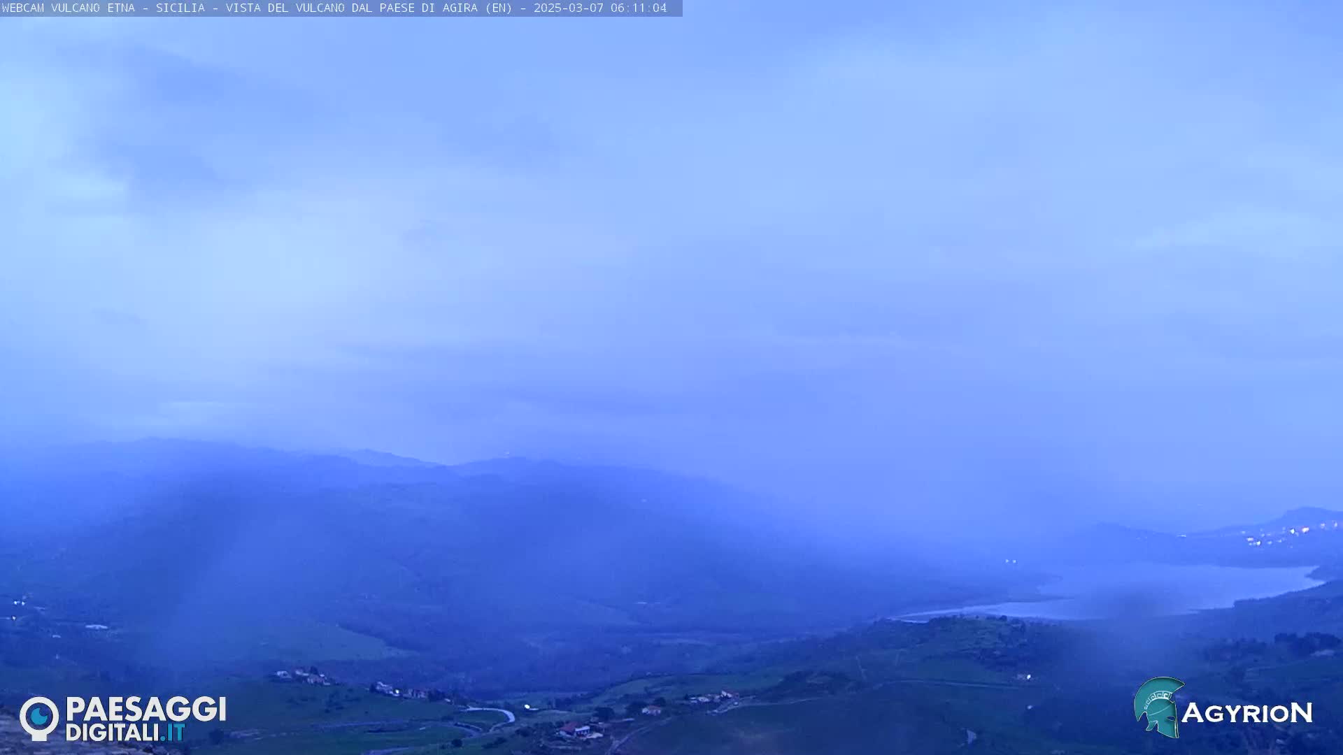 A hazy, bluish-purple dawn reveals a vast, rolling landscape of distant mountains and a valley with a body of water, under a mostly clear sky.