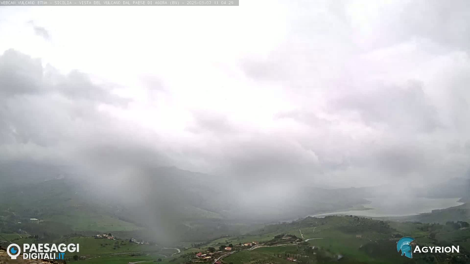 A rain-swept view of a hilly landscape with a body of water in the distance, under a cloudy sky.