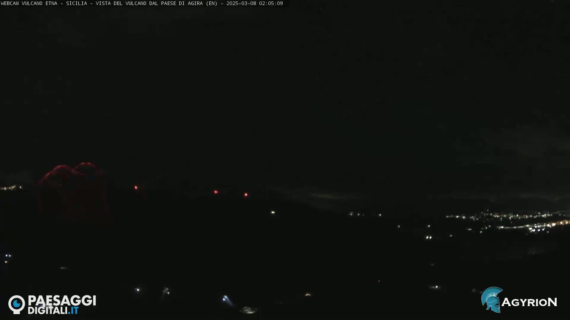 A nighttime, long shot of a dark landscape with a faintly glowing red volcano in the distance, and a few scattered lights below.