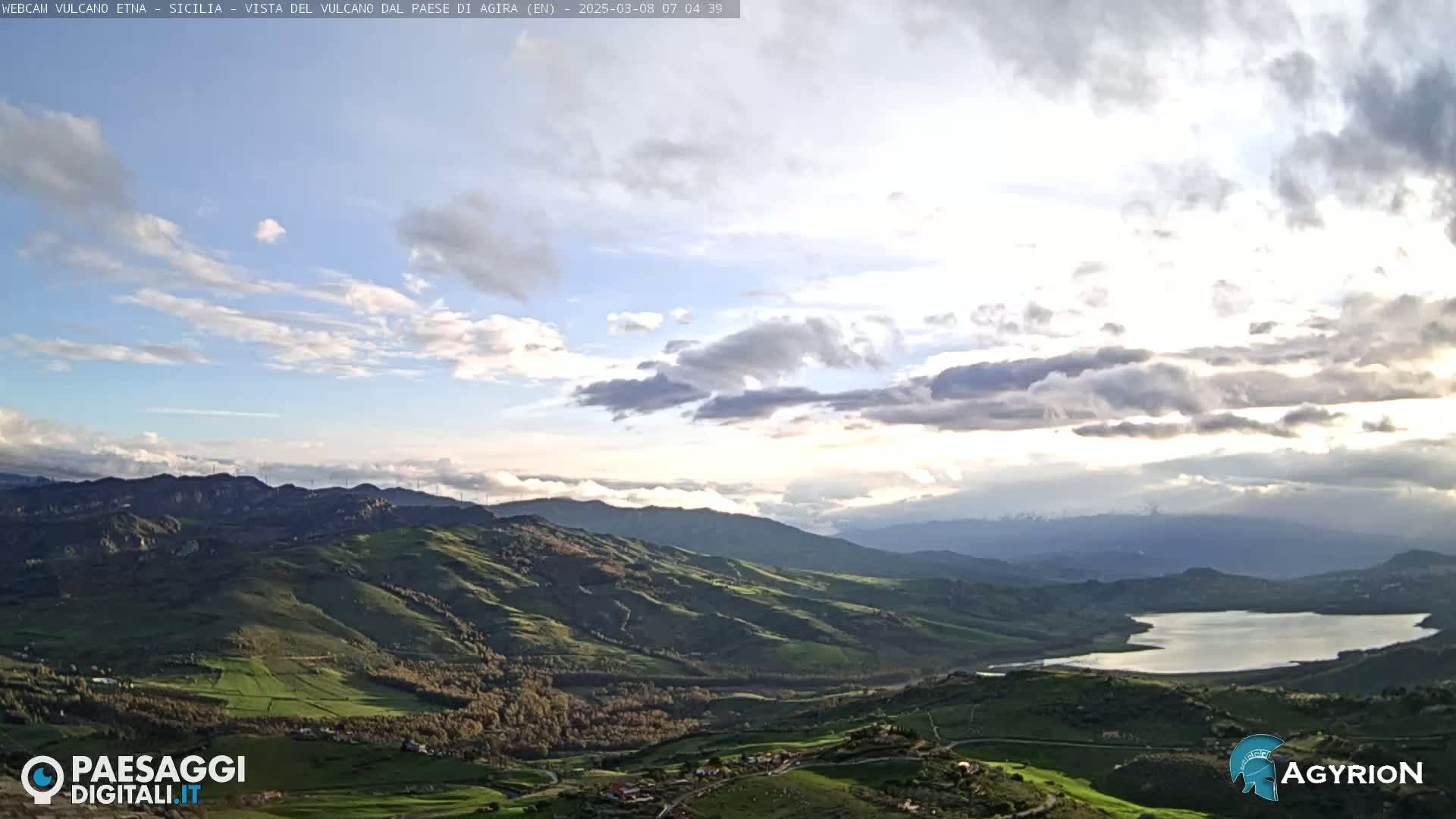 A partly cloudy sky overlooks a vast, rolling green landscape with a lake nestled in a valley.