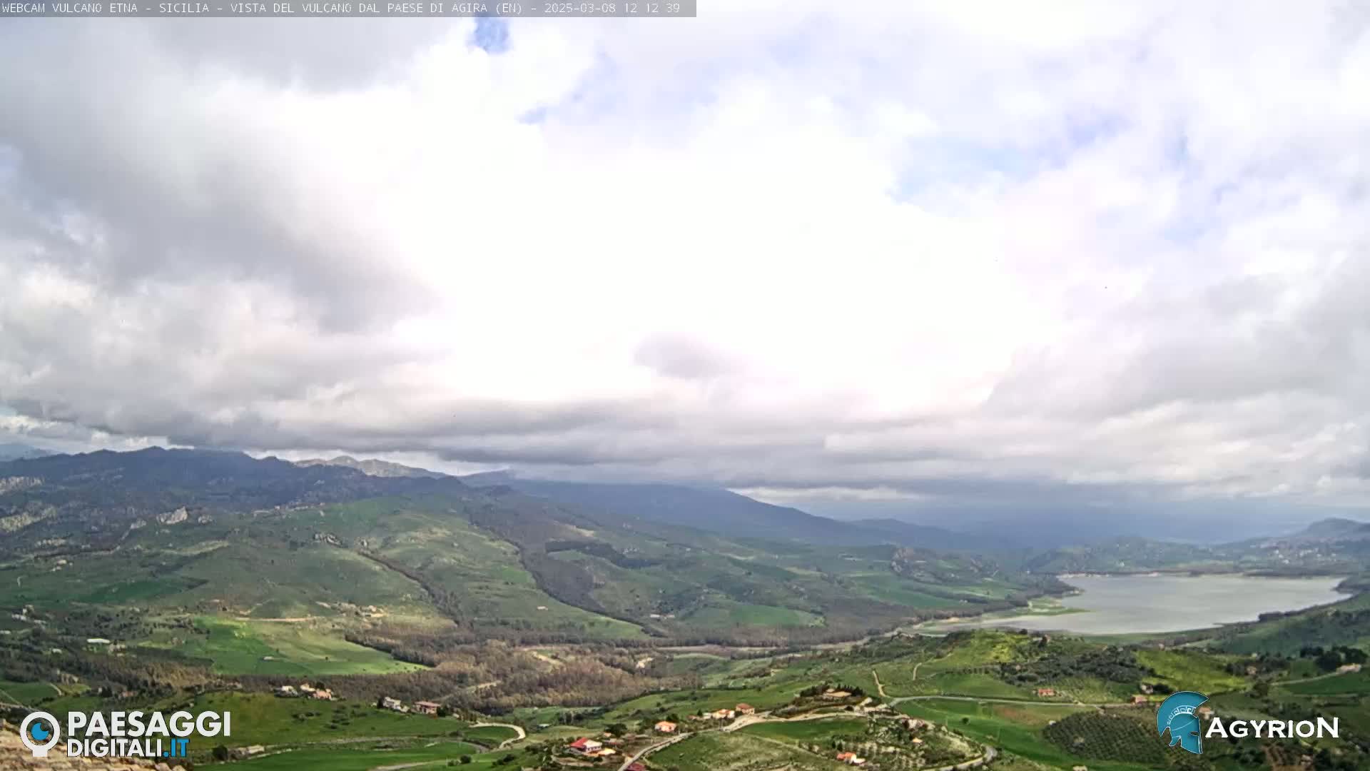 A partly cloudy day reveals a sweeping vista of rolling green hills and a lake nestled in a valley, with mountains in the distance.