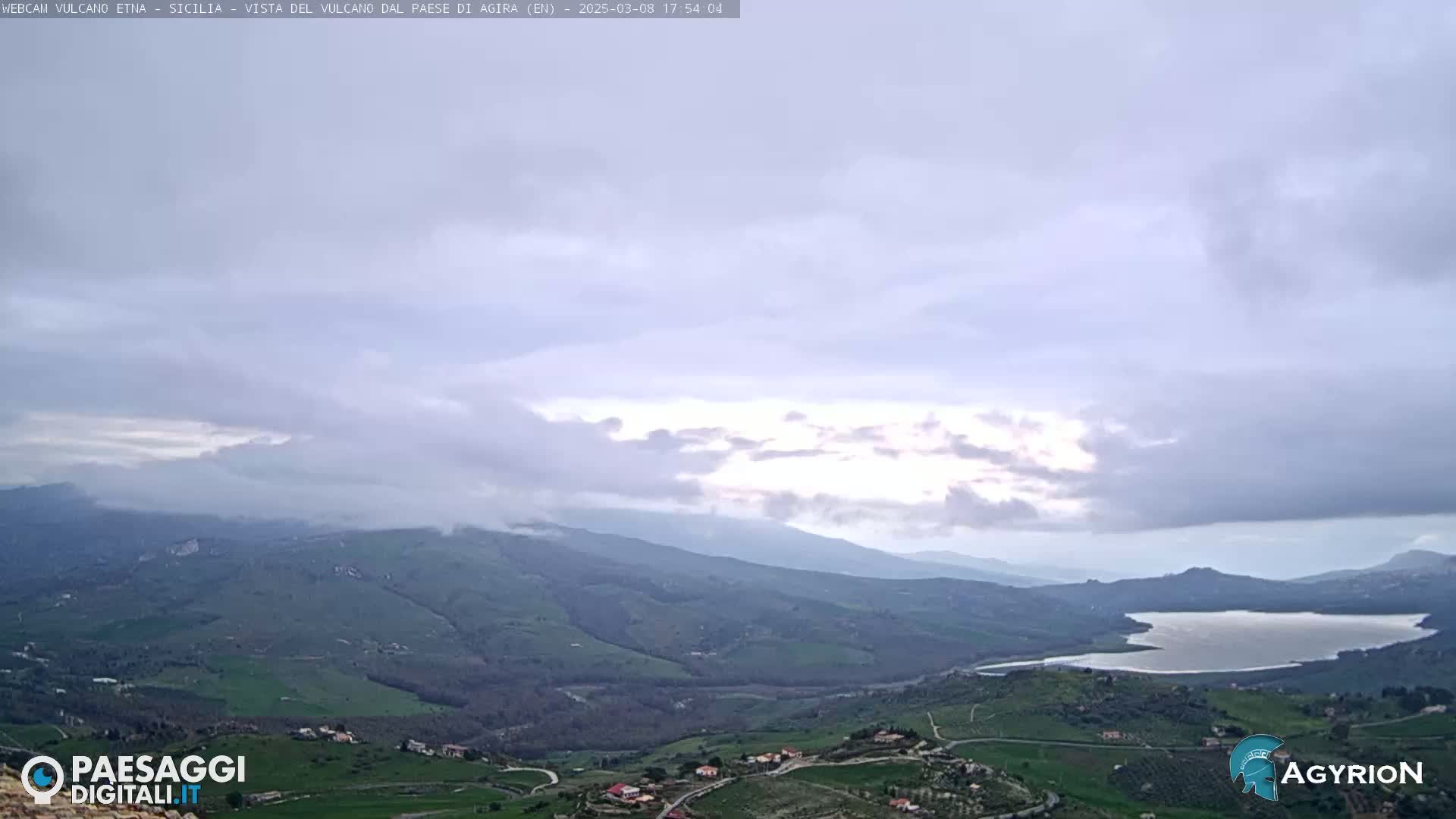 A vast, hilly landscape with a lake stretches out under a mostly cloudy sky, with some brighter areas breaking through the clouds.