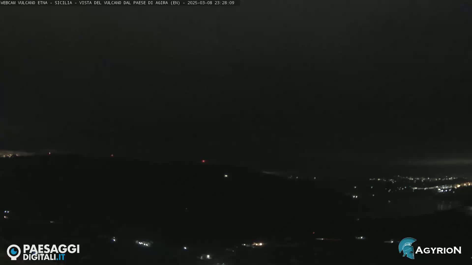 A nighttime, dark view of a hilly landscape with scattered lights from a distant town under a mostly clear sky.