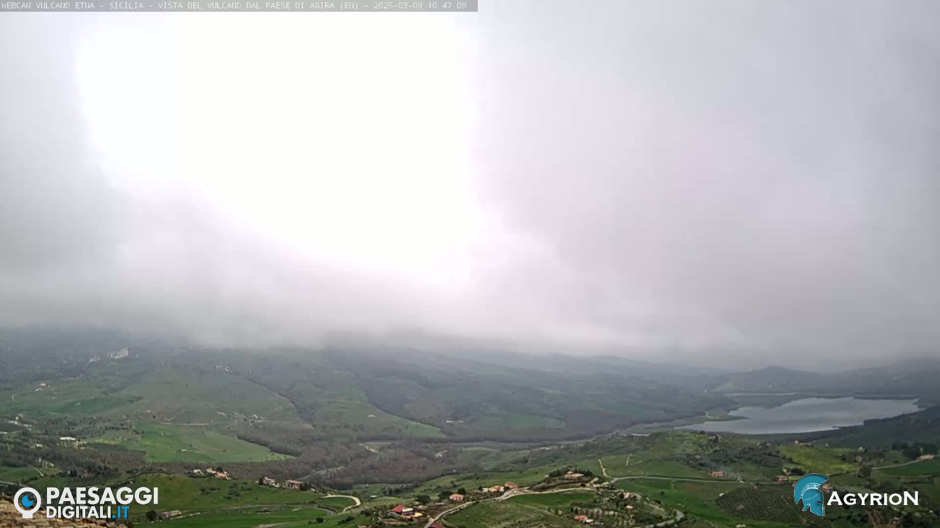 A foggy, overcast day reveals a hilly landscape with a lake in the distance and scattered farmhouses nestled in the valleys.