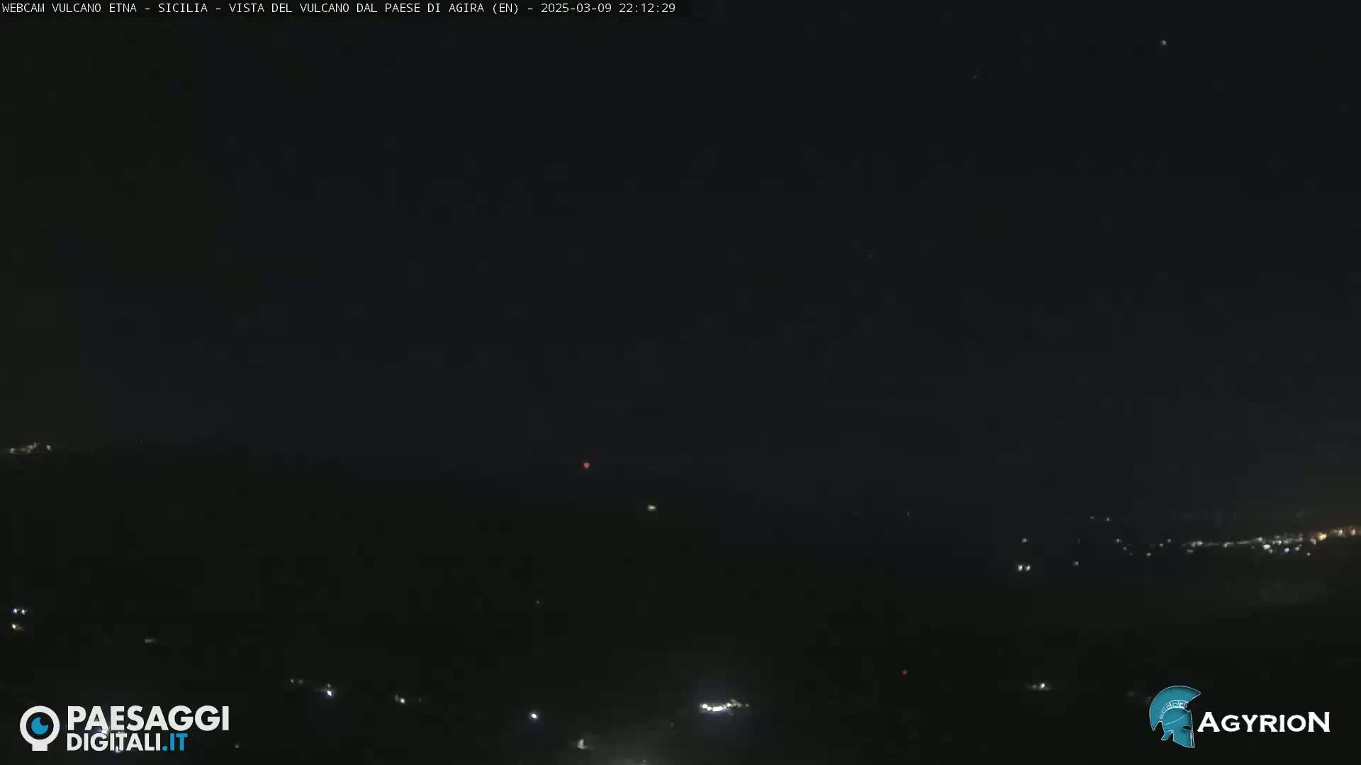 A nighttime long shot of a dark landscape with a few scattered lights in the distance under a clear, starlit sky.