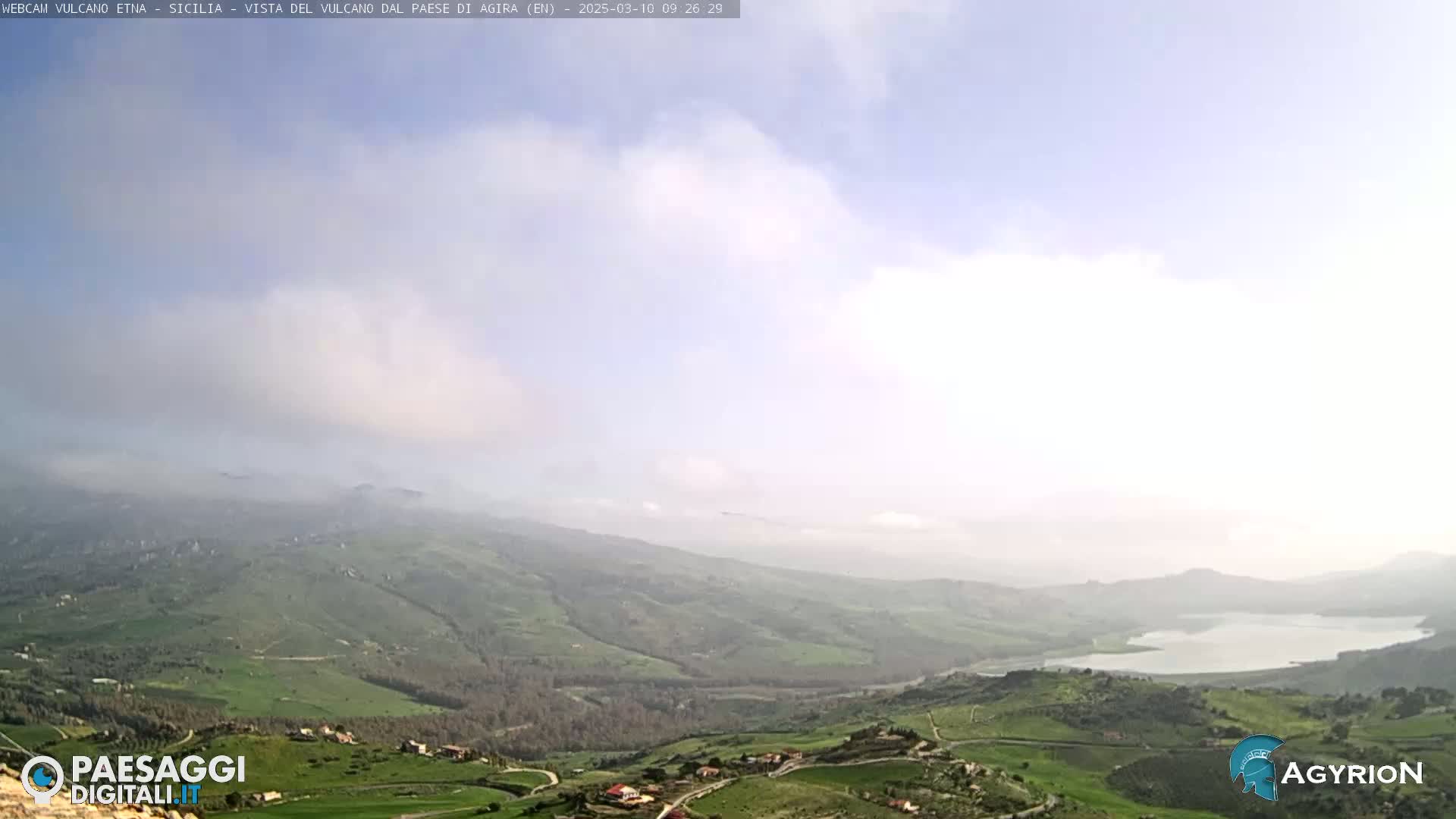 A hazy, partly cloudy day reveals a panoramic view of a rolling green landscape with a lake in the distance.