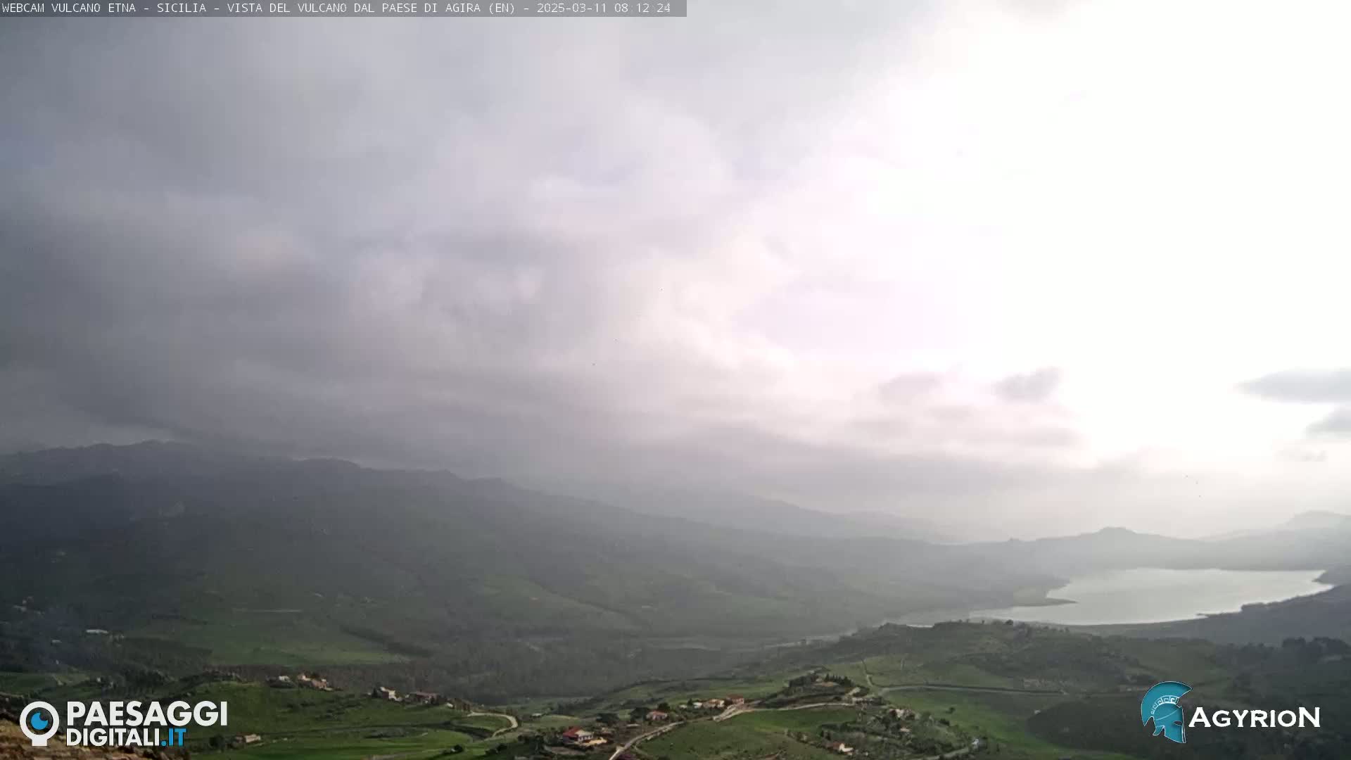 A hazy, overcast sky overlooks a vast, hilly landscape dotted with small settlements and a lake in the distance.